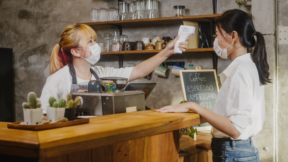 young-female-restaurant-staff-wearing-protective-face-mask-using-infrared-thermometer-checker-or-temperature-gun-on-customer-s-forehead-before-enter